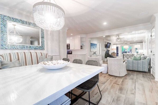 A spacious living area with a large dining table, a chandelier, and a modern kitchen in the background.