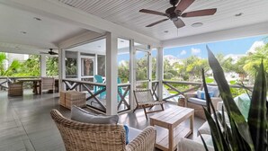 Un espacio interior amplio con mobiliario de mimbre, ventilador de techo y grandes ventanales con vistas a una piscina y jardines.