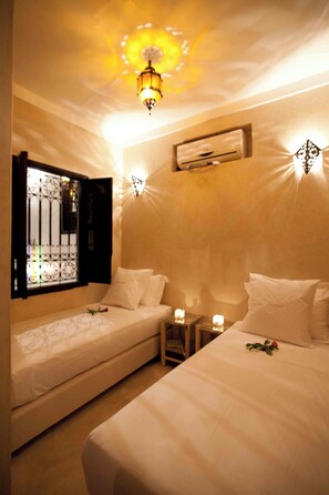 A bedroom with two beds, a window with a decorative metal grill, a wall-mounted air conditioner, and a ceiling light fixture.