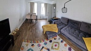 Uma sala de estar com sofá, televisão, área de jantar com cadeiras e cozinha com mesa e balcão.