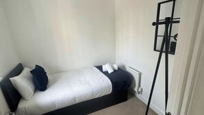 Uma cama de solteiro com estrutura preta, roupa de cama branca e cabeceira cinza. Há um suporte de metal preto com um espelho e uma pequena prateleira encostada na parede.
