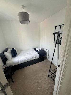 Un lit simple avec un cadre noir, une literie blanche et une tête de lit grise. Contre le mur, on trouve un présentoir en métal noir avec un miroir et une petite étagère.