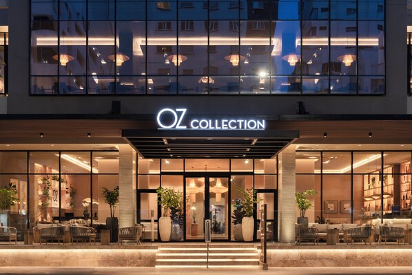Entrada moderna de hotel con fachada de vidrio, zona de estar al aire libre y un letrero que dice "Oz Collection".