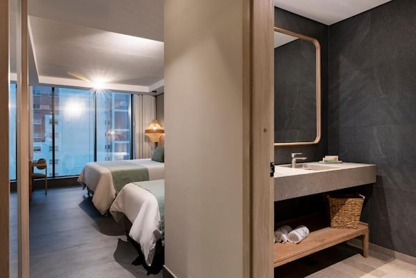 A modern hotel room with a large bed, a vanity with a mirror, and a view of the city through large windows.