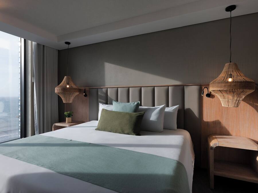 Une chambre d’hôtel moderne dotée d’un grand lit, de deux tables de chevet, d’une chaise en bois et d’une grande fenêtre donnant sur la ville.