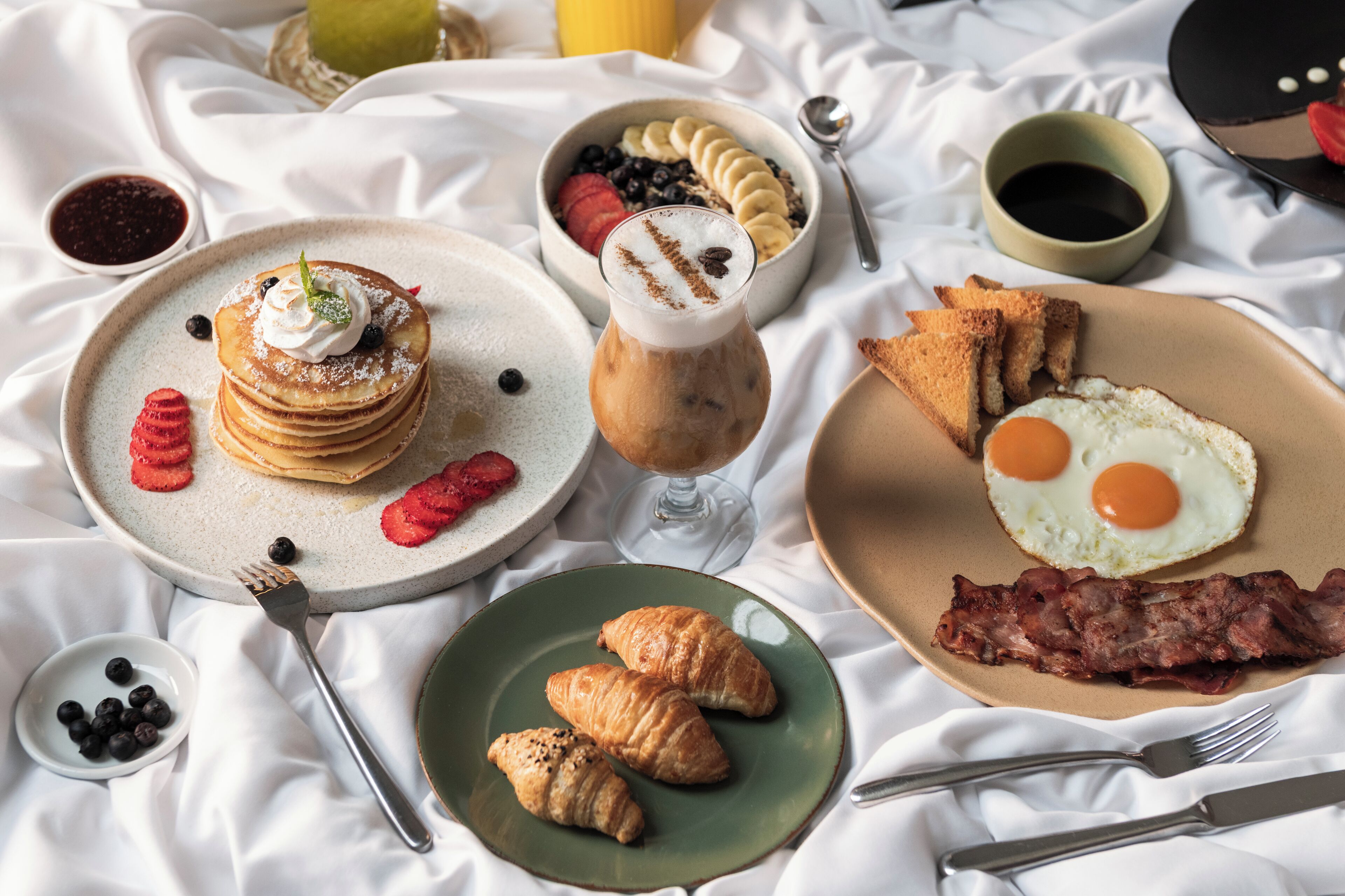 A breakfast setting with pancakes, eggs, croissants, and a coffee drink on a white cloth.