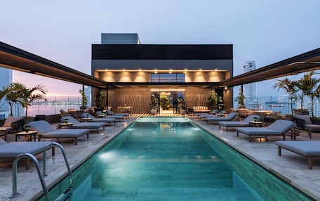 A rooftop pool area with lounge chairs and a bar, overlooking a cityscape.
