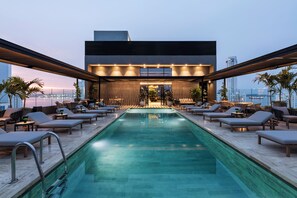 A rooftop pool area with lounge chairs and a bar, overlooking a cityscape.