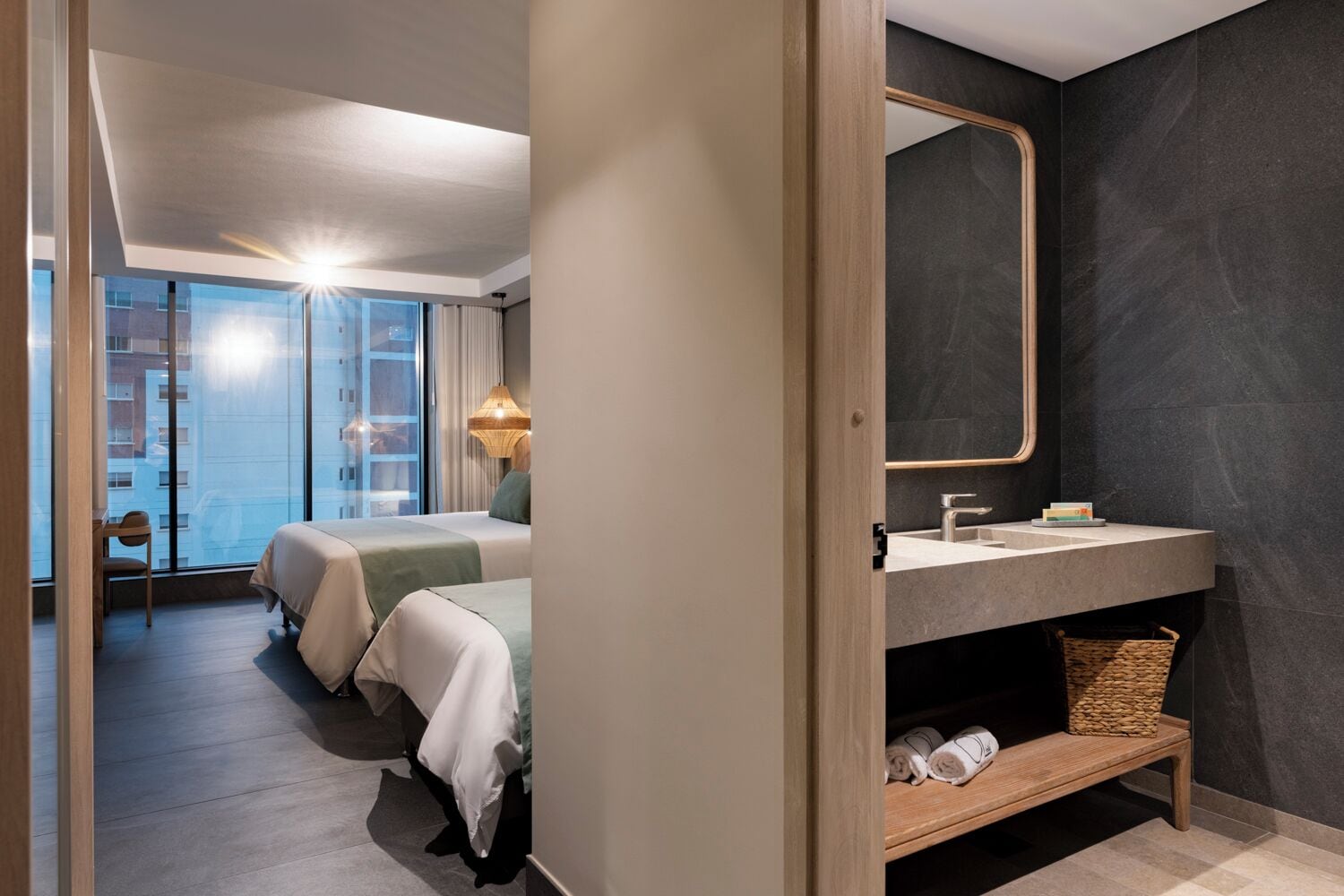 Una habitación de hotel moderna con una cama grande, un baño con pared oscura y un espejo, y vistas de la ciudad a través de amplios ventanales.
