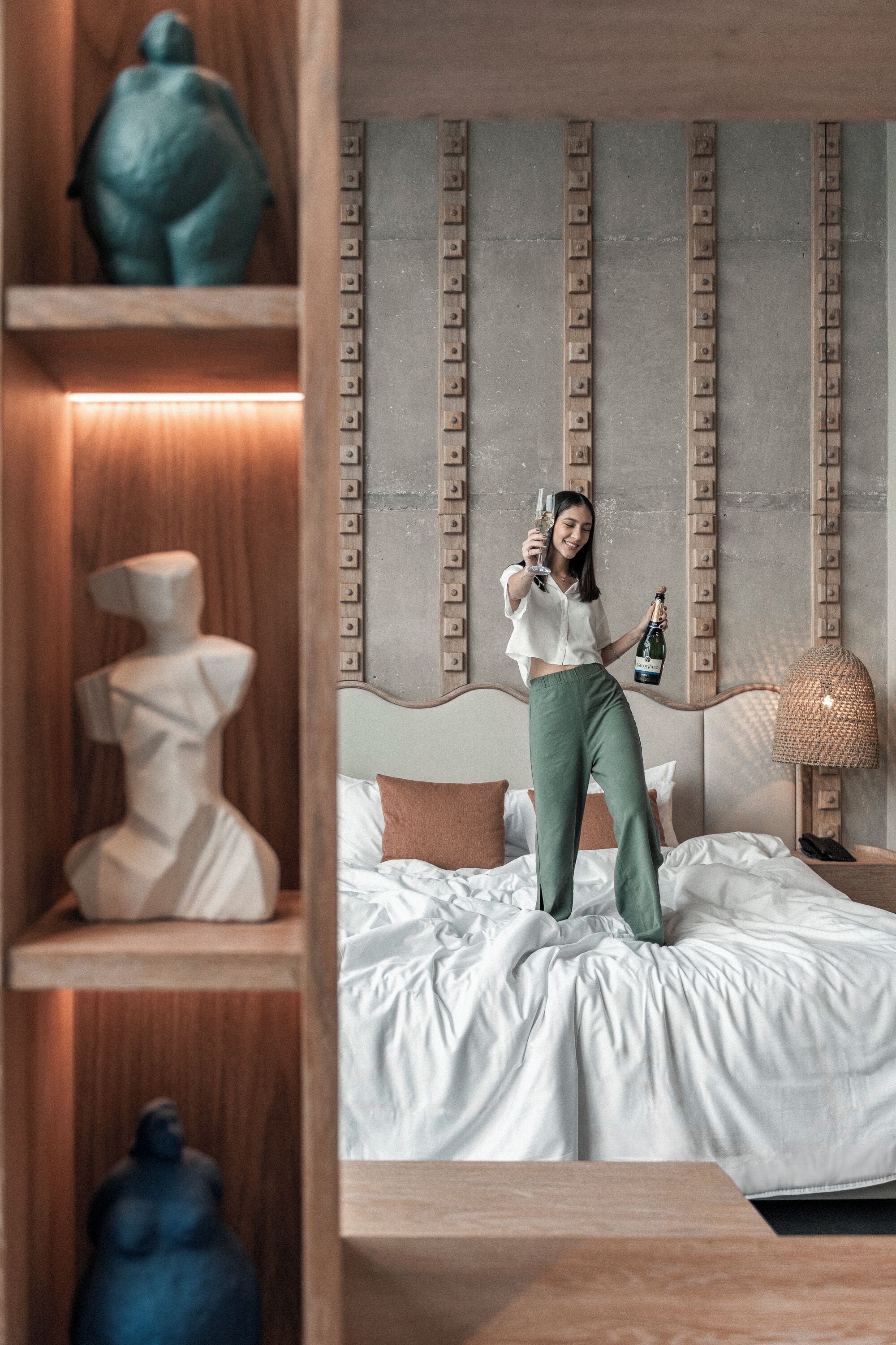 A person standing on a bed in a room with a modern design, including a headboard with vertical lines and a wooden shelf with decorative items.