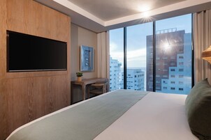A modern hotel room with a large bed, a desk, a flat-screen TV, and a city view.