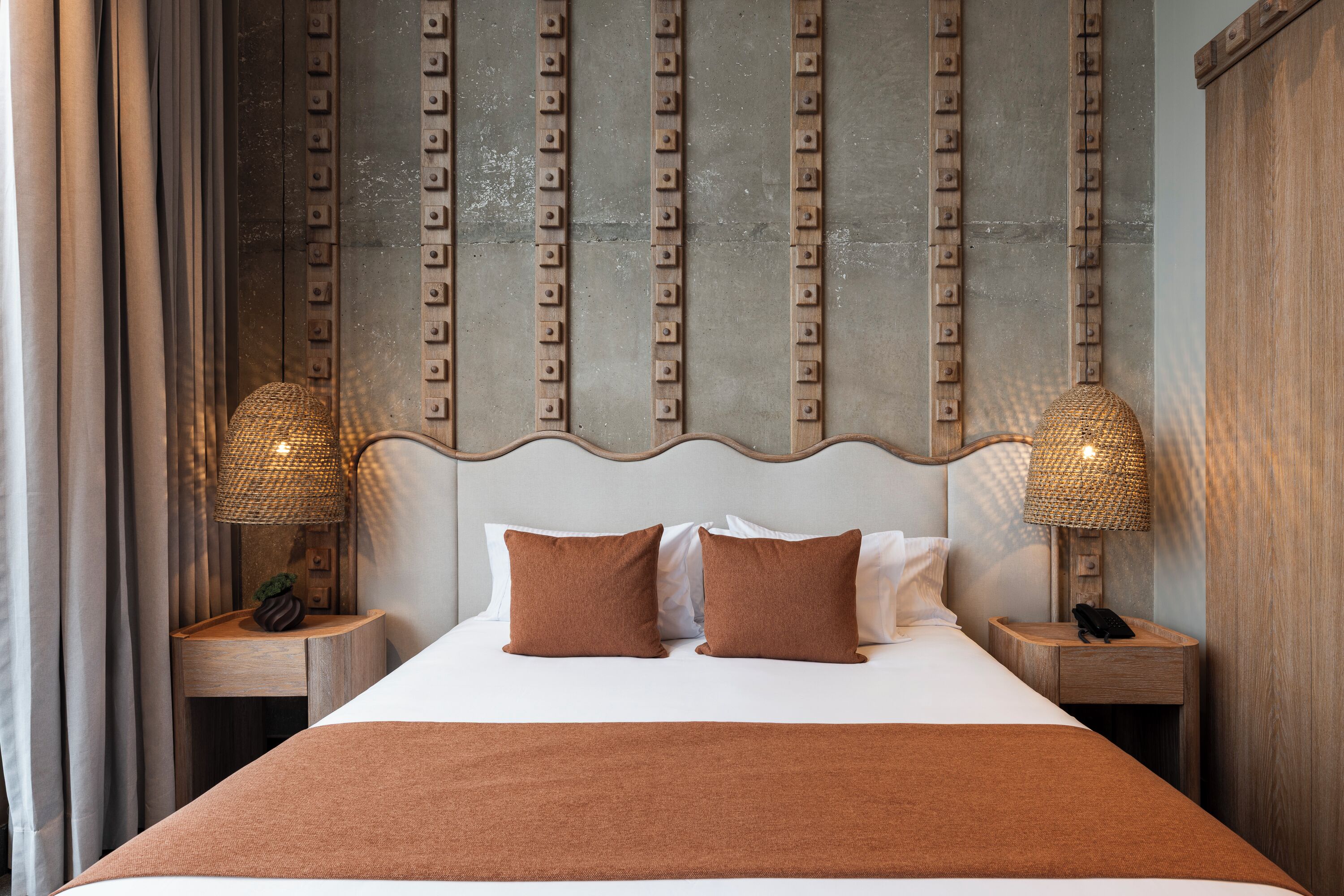 A neatly made bed with a brown throw blanket, two brown pillows, and a headboard with vertical metal studs. There are two bedside tables with lamps and a phone on one of them.