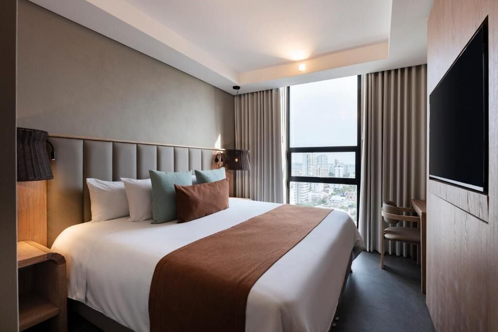 A modern hotel room with a large bed, a flat-screen TV, a desk, and a view of the city through large windows.