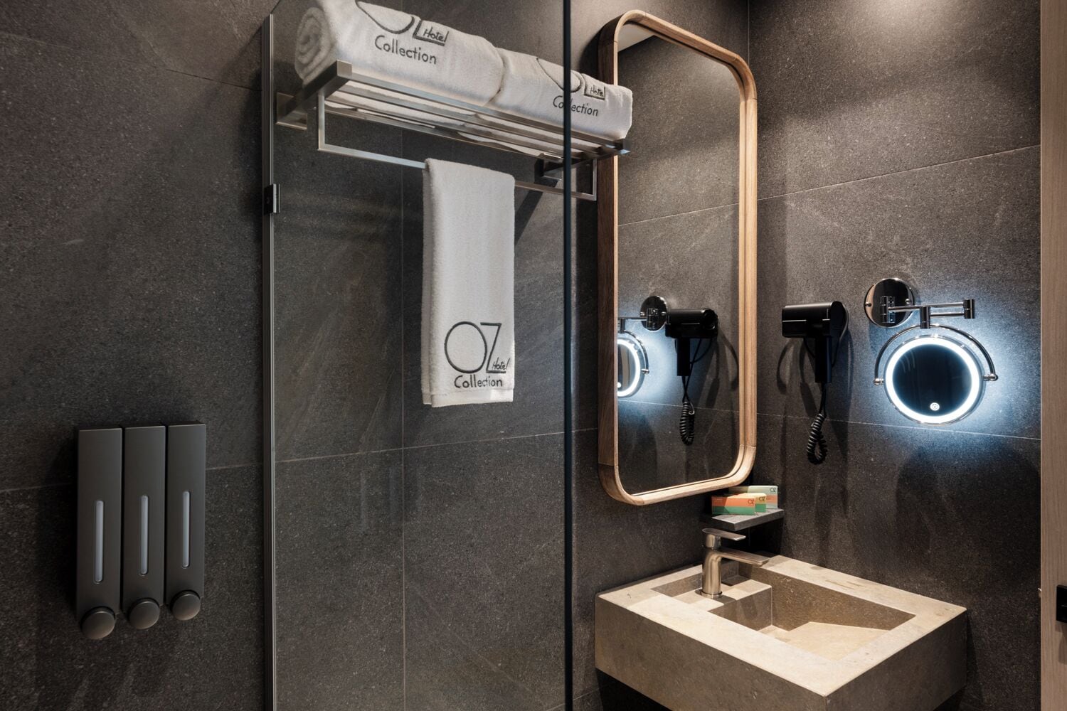 A modern bathroom with a glass shower enclosure, a towel rack with a hotel towel, a mirror with a wooden frame, a sink with a faucet, and two hair dryers mounted on the wall.