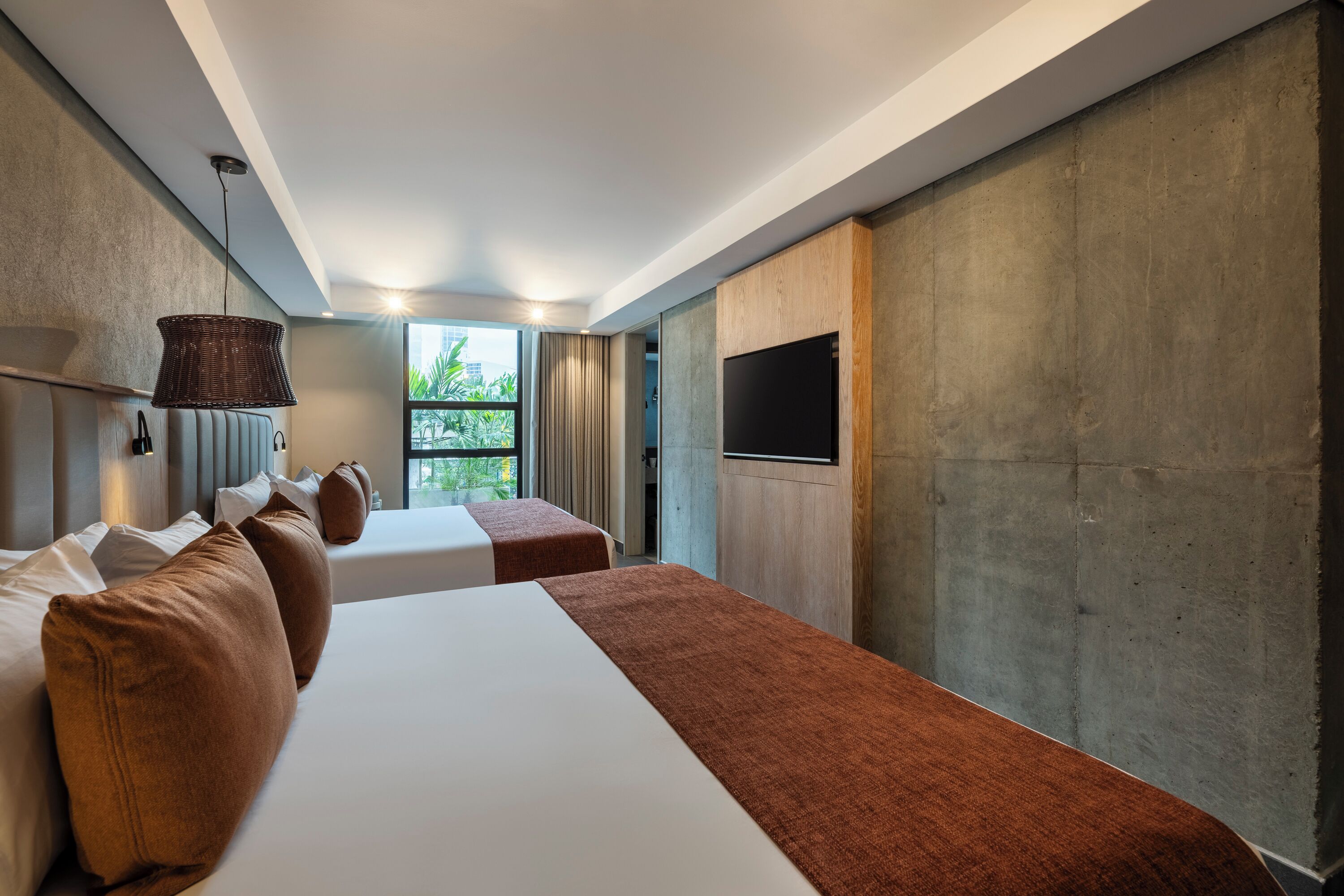 A modern hotel room with two beds, a large window, a flat-screen TV, and a concrete accent wall.