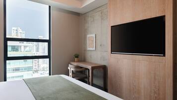 Una habitación de hotel moderna con una cama grande, un escritorio, televisión y vistas de la ciudad a través de amplios ventanales.