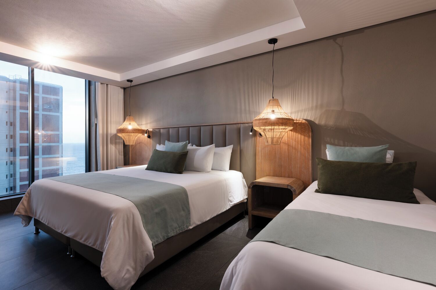 Habitación de hotel con dos camas, un ventanal con vistas a edificios y dos lámparas fijadas a la pared.