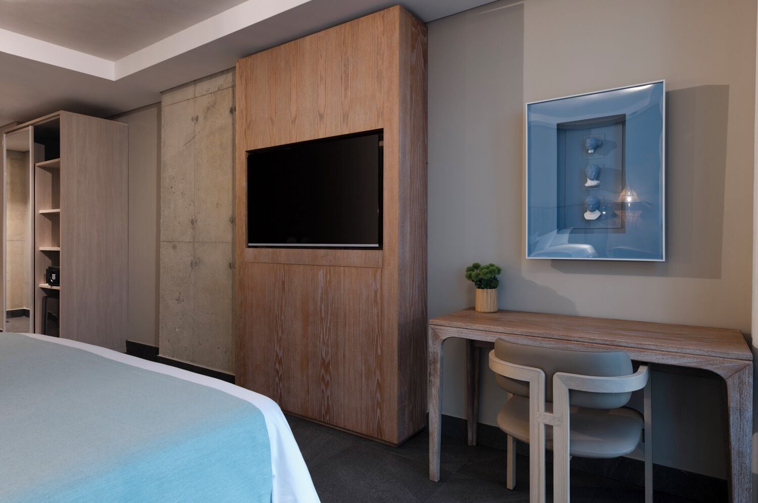 A modern hotel room with a bed, a wooden wardrobe, a desk with a chair, and a television mounted on the wall.