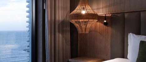 A hotel room with a large window, a bed with white linens, a wooden nightstand, and a woven pendant light fixture.