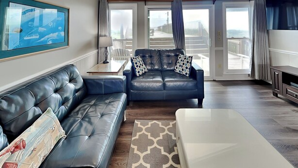 Ein modernes Wohnzimmer mit einer schwarzen Ledersofa, einem weißen Couchtisch und einem blauen Sessel mit gemusterten Kissen. Ein gerahmtes Bild hängt an der Wand, und ein großes Fenster bietet einen Ausblick ins Freie.