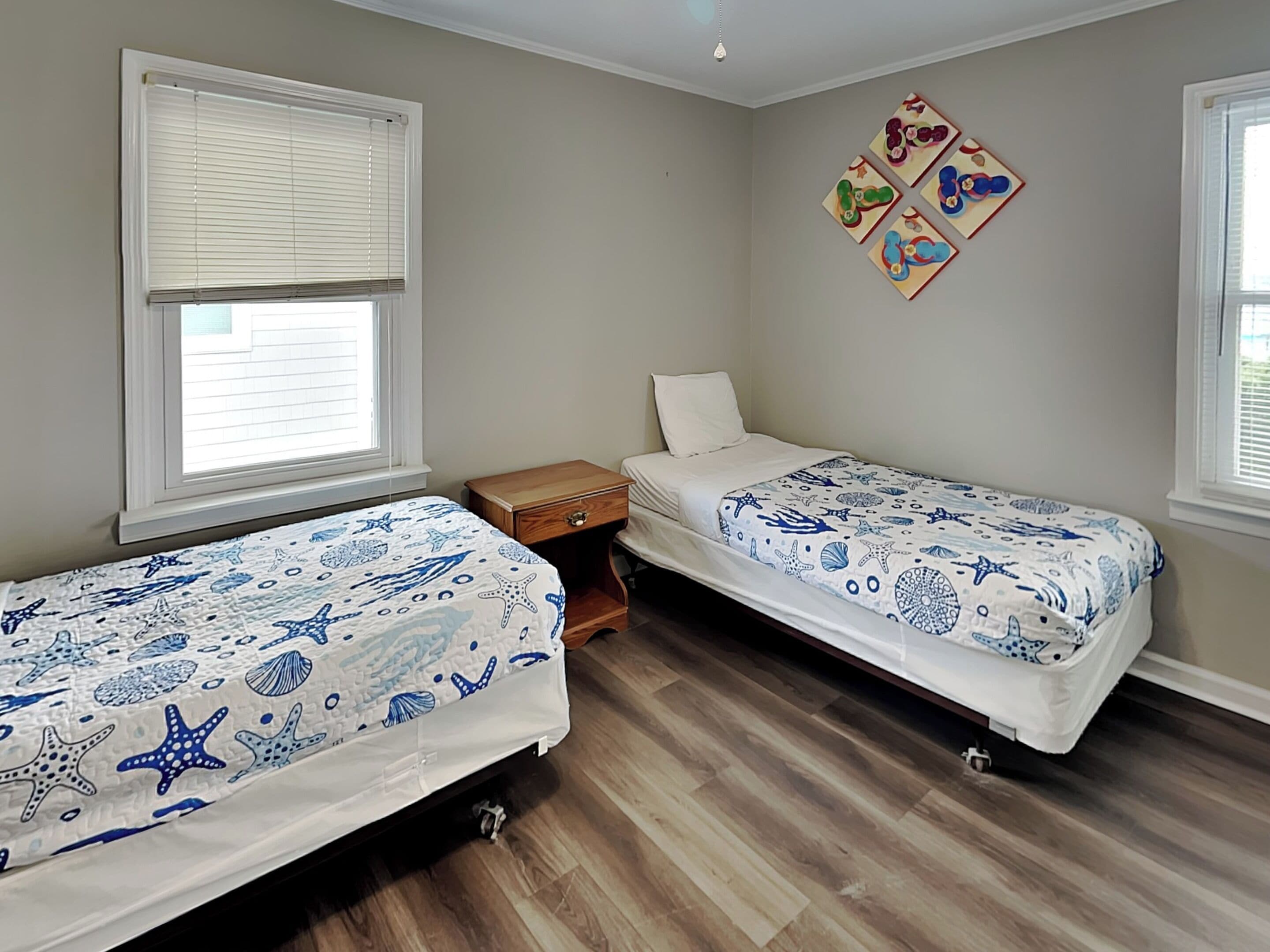 Una camera con due letti, ognuno con una coperta a tema oceano in blu e bianco, un comodino in legno e una parete decorata con opere d'arte colorate.
