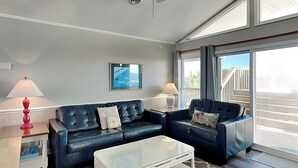 Ein modernes Wohnzimmer mit einem schwarzen Ledersofa, einem weißen Couchtisch und einem Tisch mit Glasplatte. Auf Beistelltischen stehen zwei Lampen, an der Wand hängt ein gerahmtes Bild und durch große Fenster ist ein Ausblick ins Freie zu sehen.