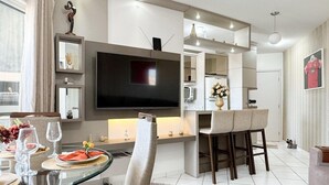 A modern living room with a glass dining table, comfortable seating, a flat-screen TV, and a kitchen area in the background. - Resid. Clube Mar Grosso-Laguna - LAG604 (Laguna)