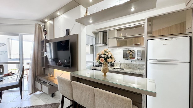A modern kitchen with a flat-screen TV, a refrigerator, and a dining area with a glass table and chairs. - Resid. Clube Mar Grosso-Laguna - LAG604 (Laguna)