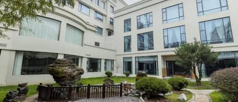 Un edificio moderno con varios pisos, grandes ventanales y un jardín ajardinado con terraza de madera y formaciones rocosas.