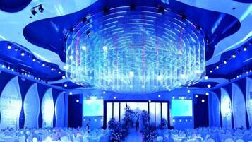 A large, modern hall with a central circular light fixture, white tables, and chairs arranged for an event.