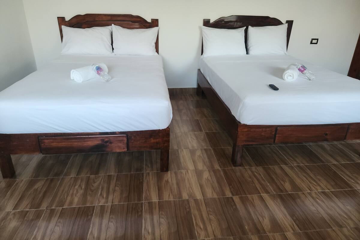 Two wooden beds with white linens and pillows, a remote control, and a rolled towel on each bed.