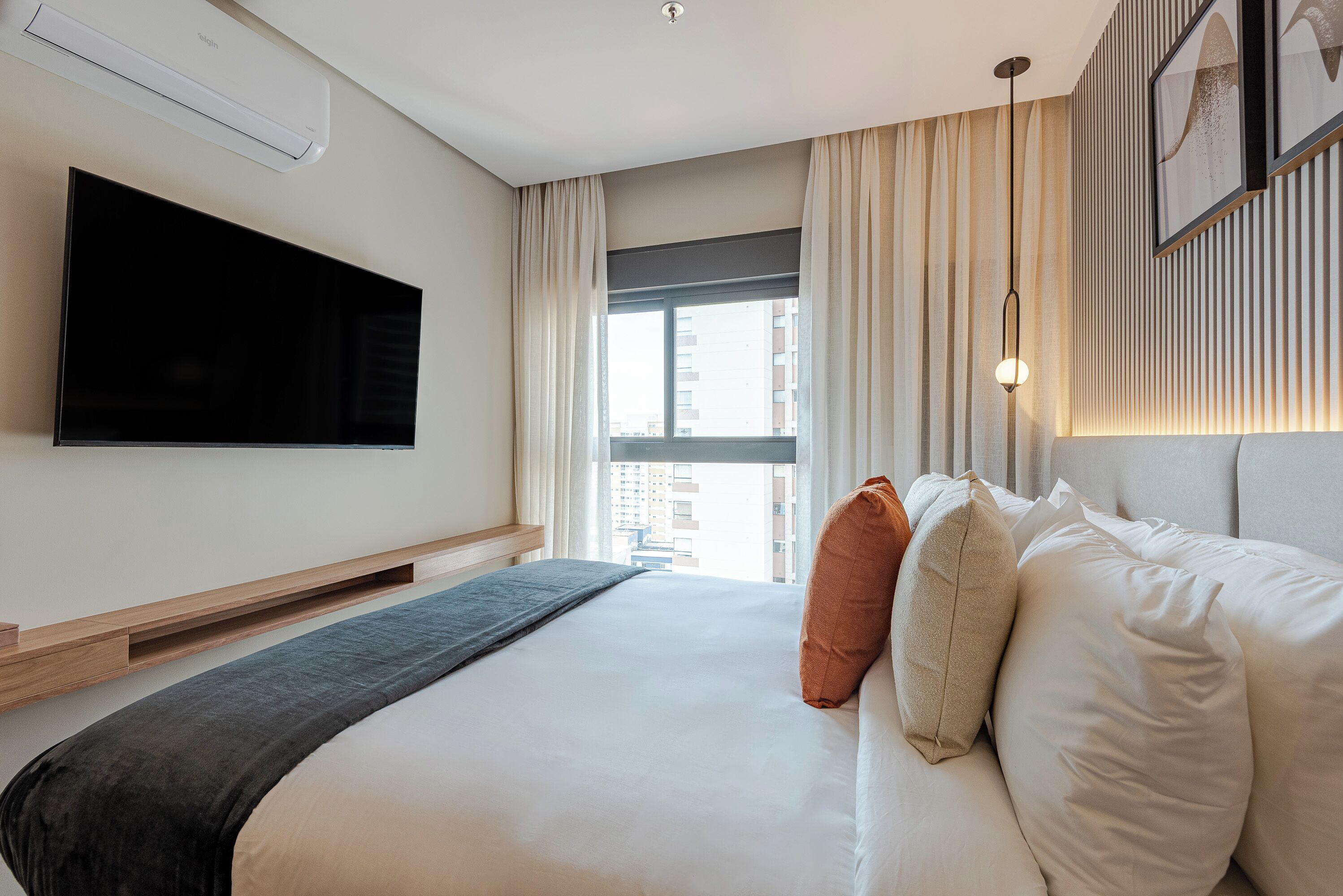 Moderni hotellihuone, jossa on suuri sänky, seinälle asennettu televisio, verhoilla varustettu ikkuna ja näkymä kaupunkiin.