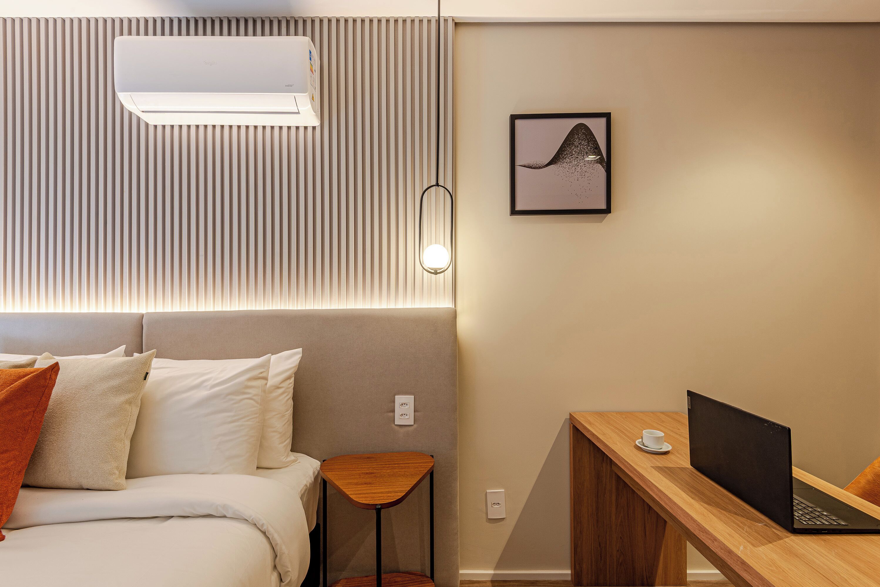 A modern hotel room with a bed, a desk with a laptop, a small table, and a wall-mounted air conditioner.