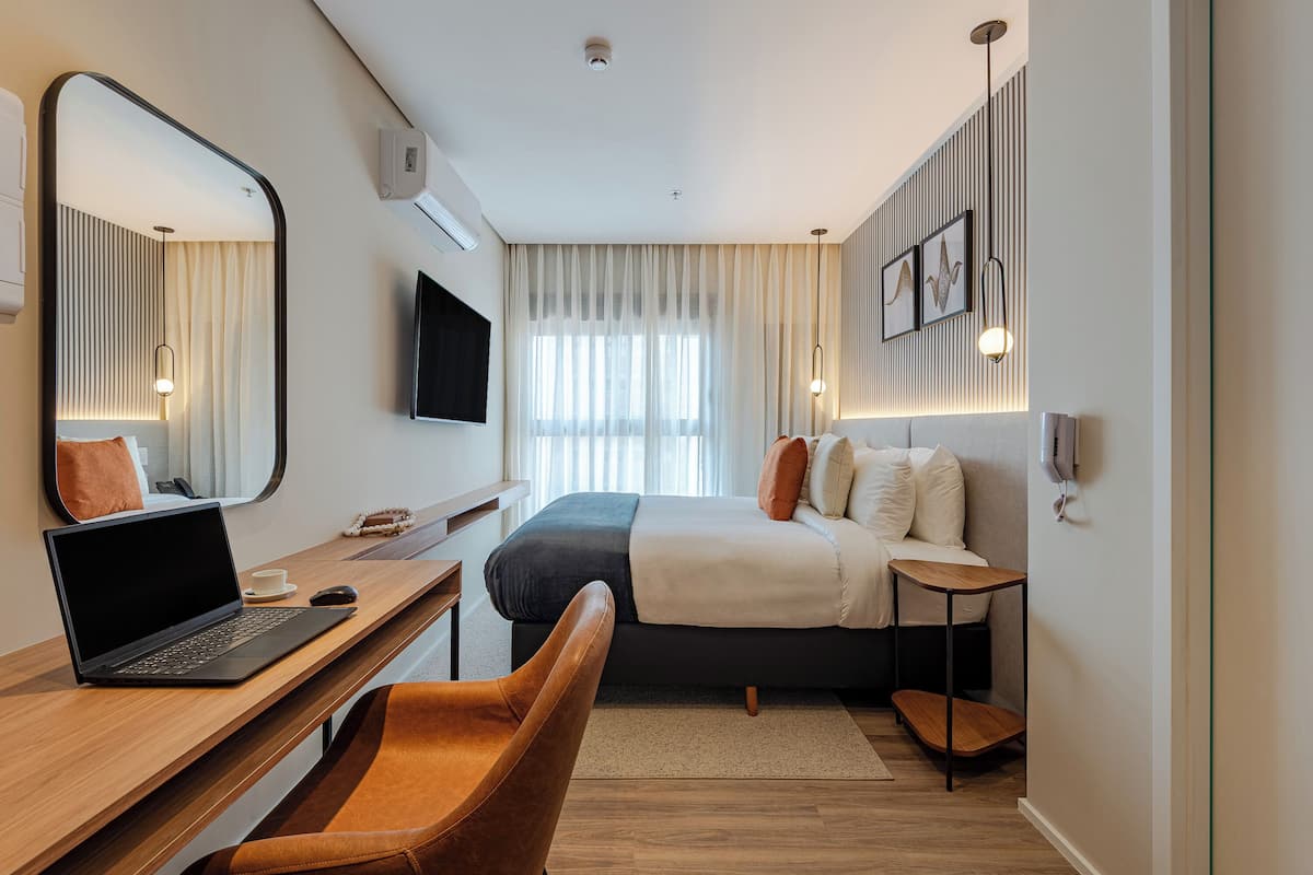 Ett modernt hotellrum med en säng, ett skrivbord med en bärbar dator, en stol, ett sängbord och en tv som är monterad på väggen.