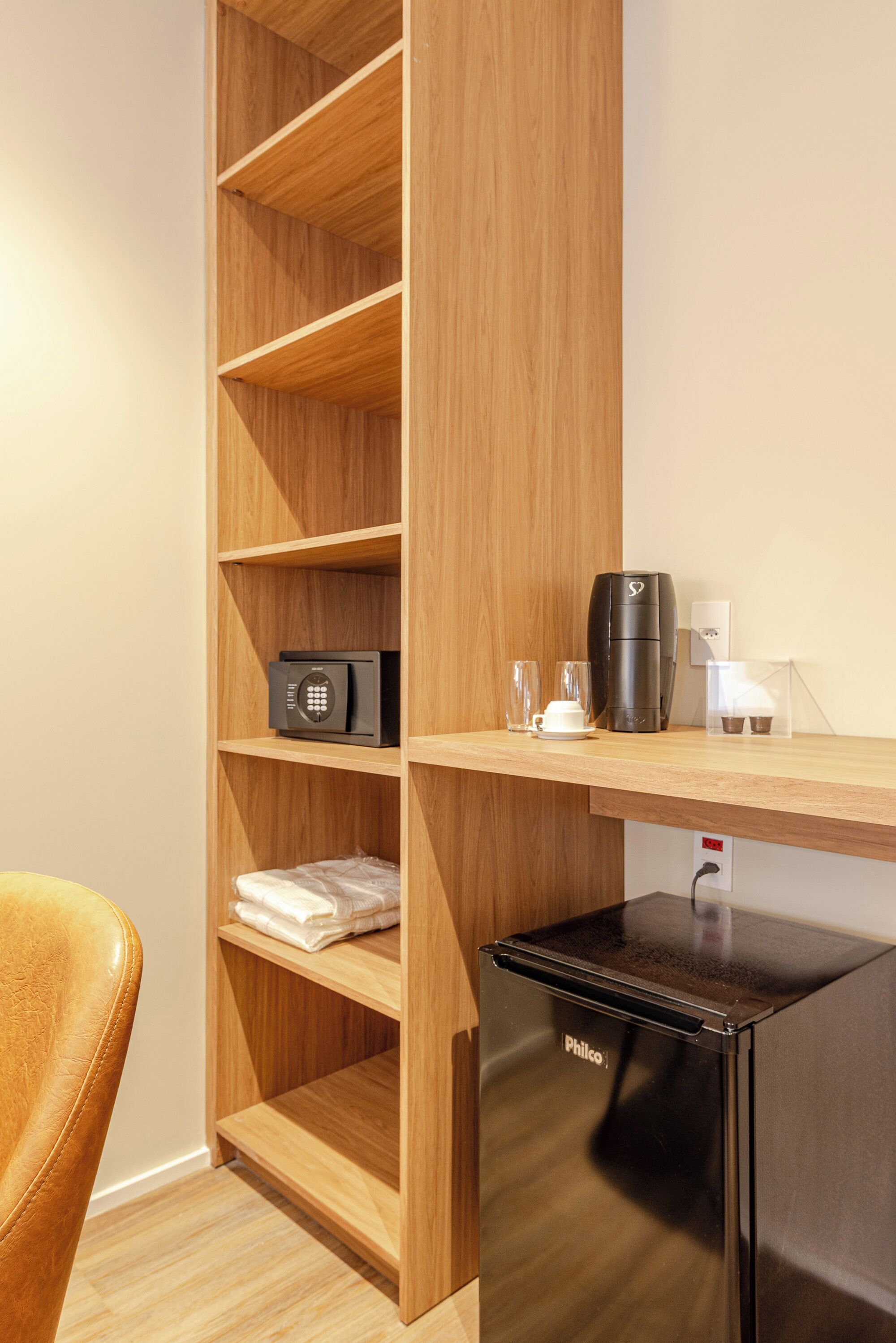 A hotel room with a wooden shelving unit, a black refrigerator, a coffee maker, and a small table with a cup and saucer.