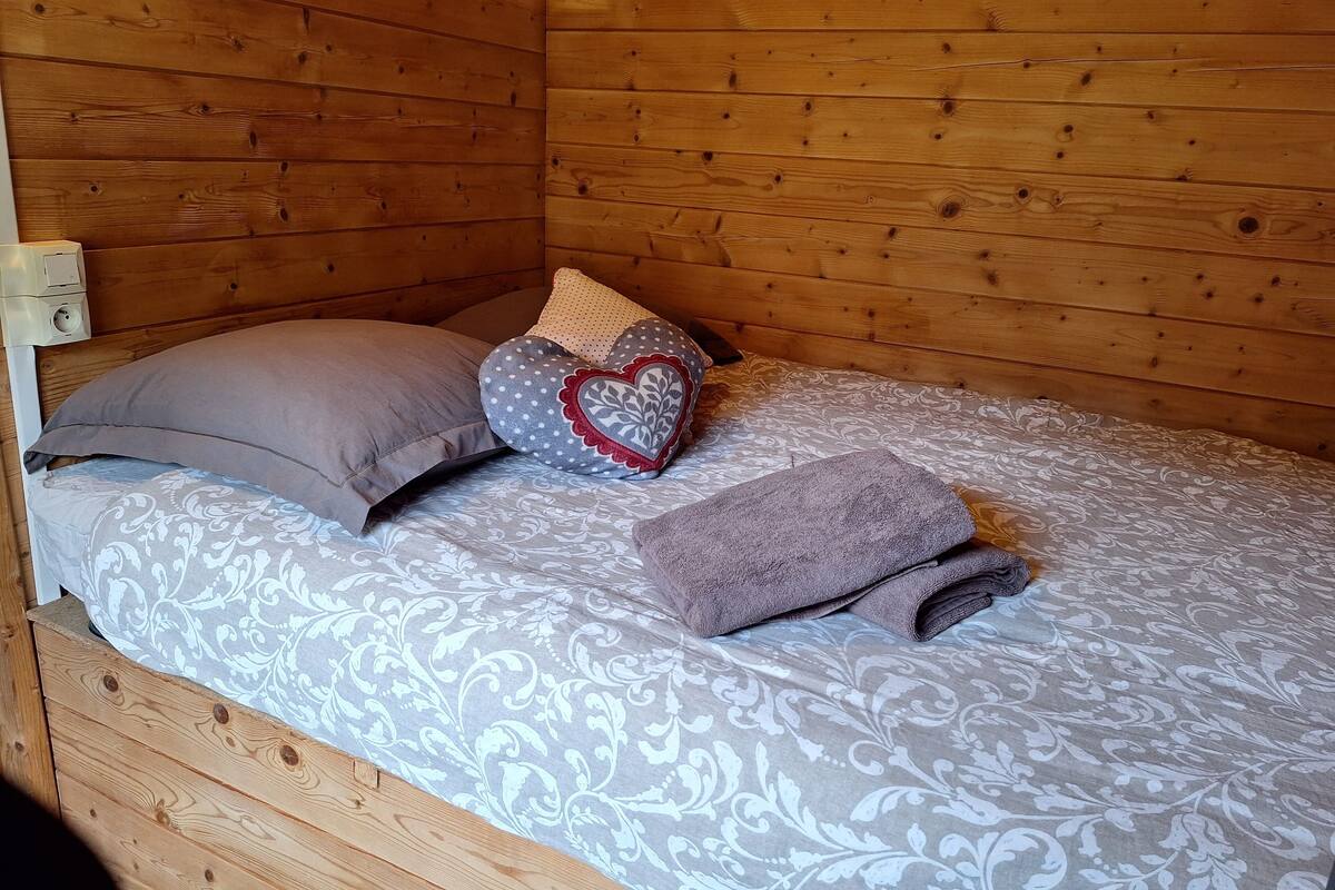 Una habitación de madera con una cama, un almohadón y un cojín con forma de corazón, además de una toalla doblada.