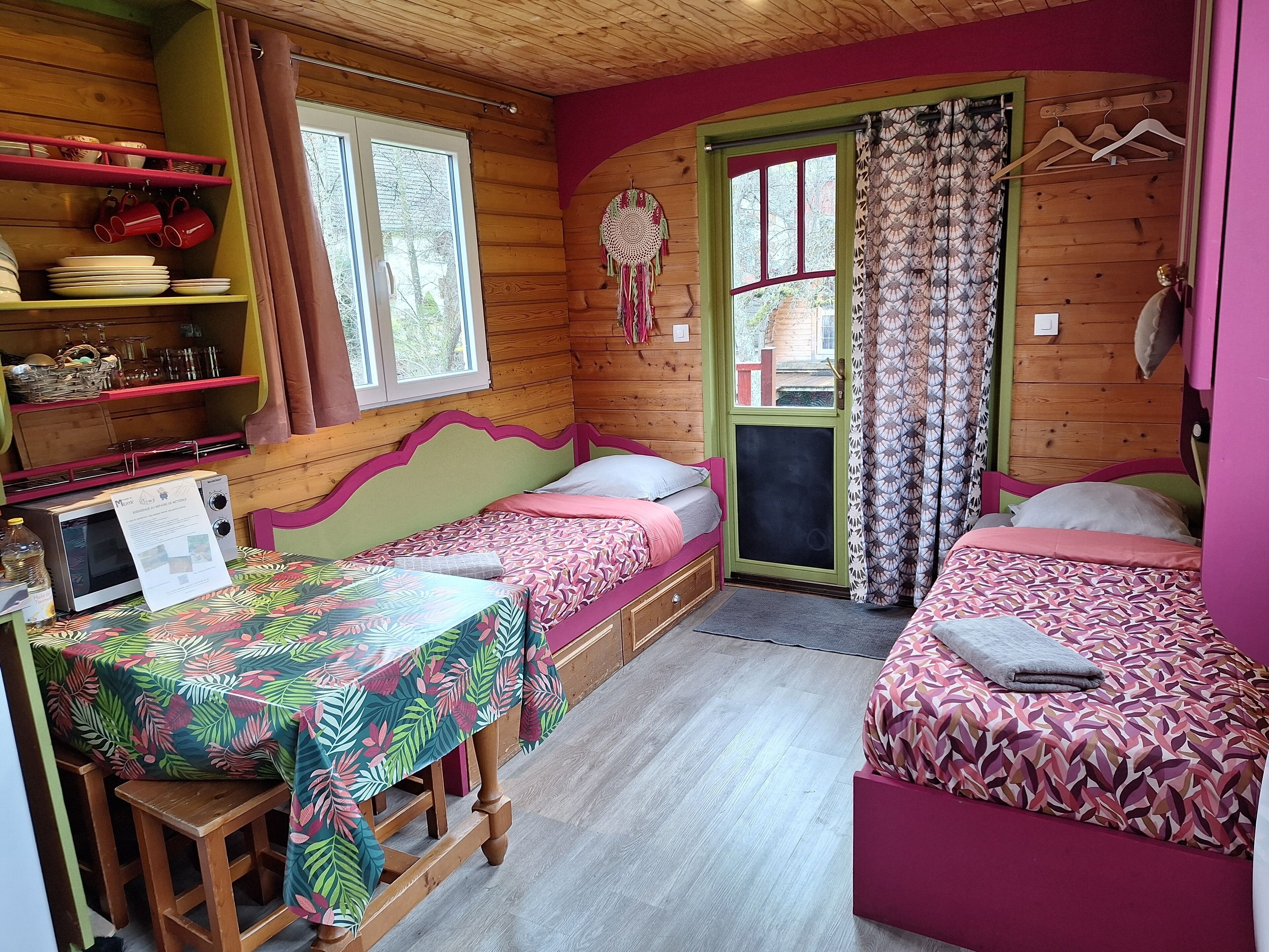 A cozy cabin-style room with two beds, a wooden table, and a shelf with dishes and mugs.