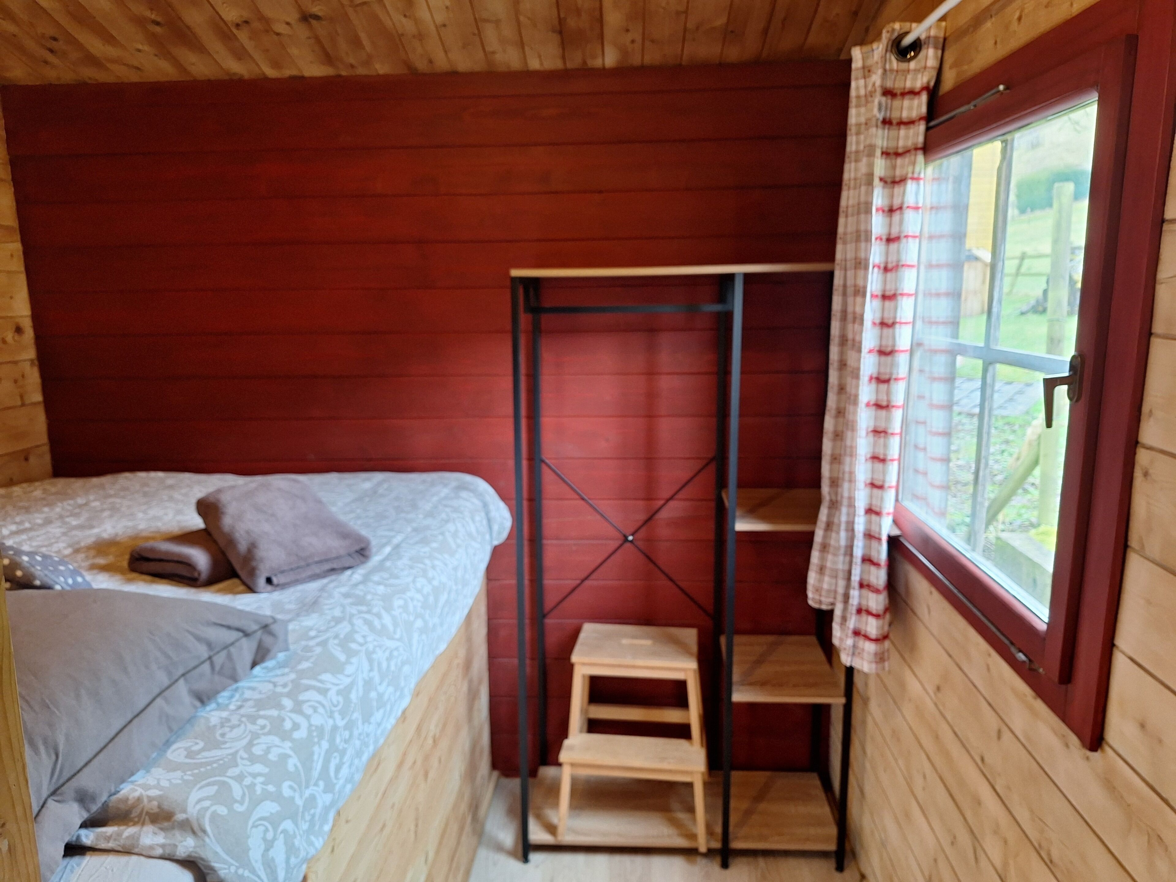 Ein Zimmer einer Holzhütte mit einem Bett, einem kleinen Holzschemel, einem Metallregal und einem Fenster mit einem karierten Vorhang.
