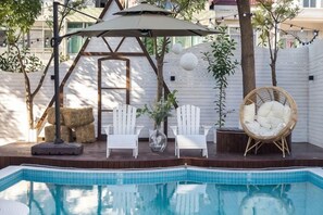 Una piscina con cubierta de madera, dos sillas Adirondack blancas, una silla de mimbre y una silla colgante bajo un toldo.