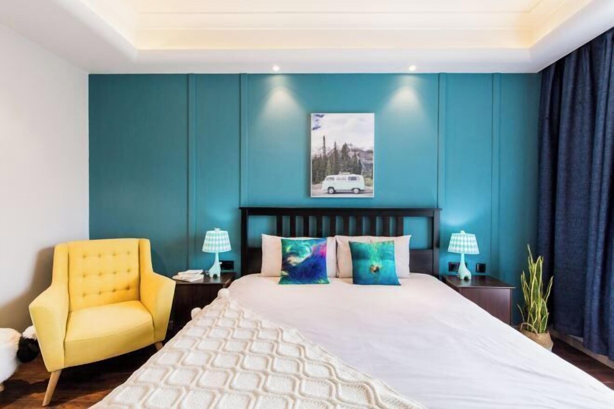 Quarto com paredes verde-água, uma cama grande com roupa de cama branca, duas mesas de cabeceira com abajures, uma poltrona amarela e um quadro de uma carrinha na parede.