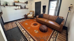 A living room with a brown leather sofa, a patterned rug, a small round ottoman, and a bookshelf with decorative items. - Cozy Cottage House in Louisville (Louisville)