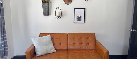 Un sofá de cuero marrón con un solo cojín, una planta de pared, una cabeza de ciervo montada, dos apliques de pared y un cuadro enmarcado.