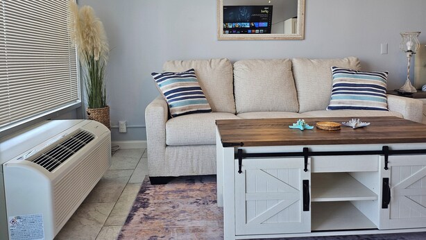 Una sala de estar con un sofá color beige, almohadas a rayas, una mesa de centro de madera, un espejo y un radiador.