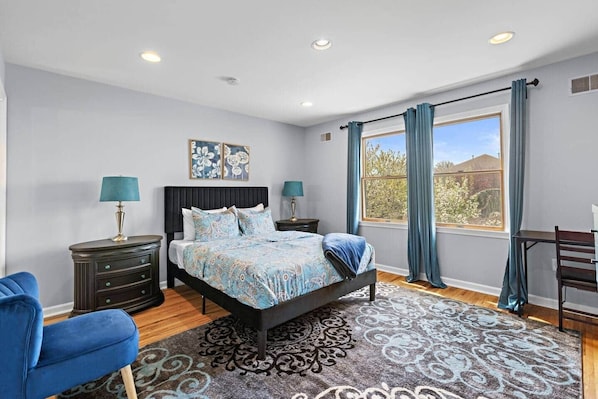 A bedroom with a bed, two bedside tables, a desk, a chair, and a large window with curtains.