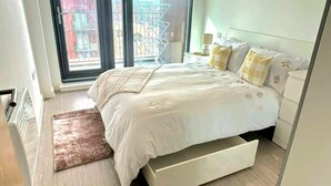 A bedroom with a bed, a nightstand, a lamp, a window with a view of buildings, and a sliding door.