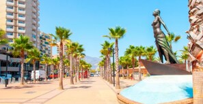 Un paseo marítimo frente a la playa con palmeras, una estatua y edificios altos al fondo.