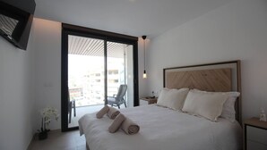 A modern hotel room with a large bed, a wooden headboard, a small bedside table, and a chair near a sliding glass door leading to an outdoor balcony. - Happy Travel Jade Tower (Fuengirola)