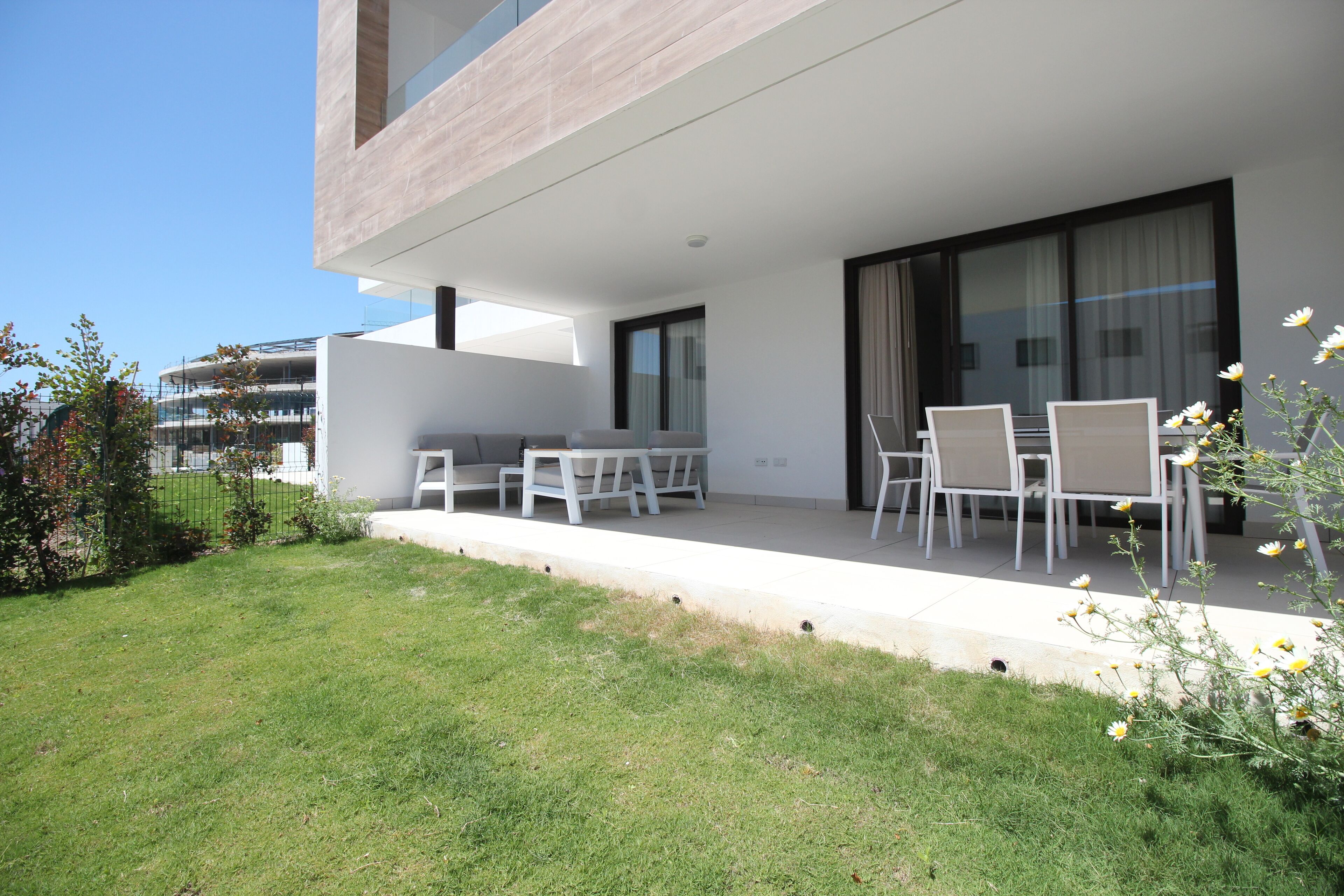 A modern outdoor patio with a sofa and dining table, surrounded by a well-maintained lawn and a small garden with flowers.