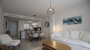 A modern living room with a white sofa, a wooden coffee table, and a dining area with chairs and a kitchen in the background. - Happy Travel Jasmine Higueron (Fuengirola)