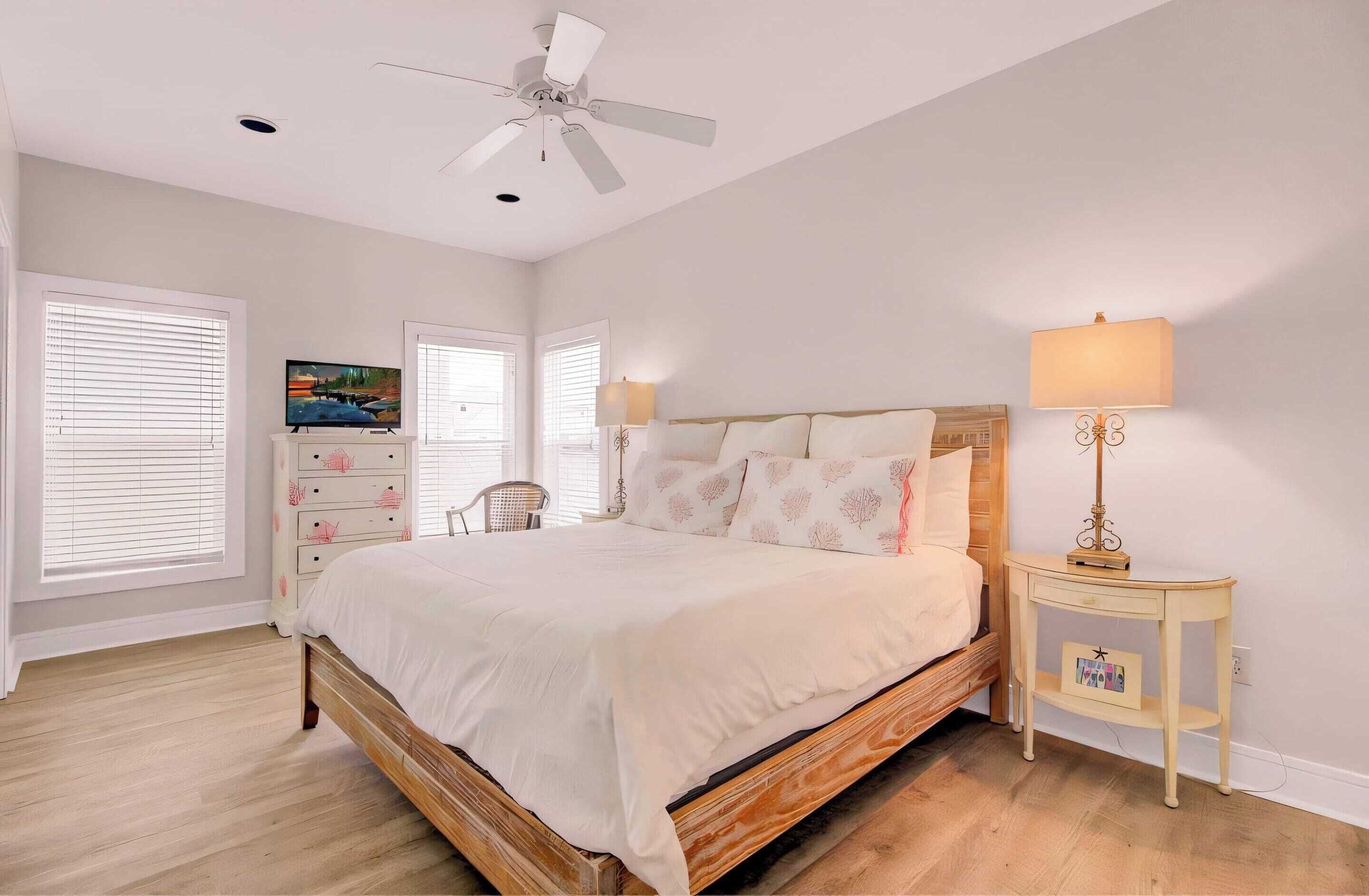 A bedroom with a wooden bed, white bedding, a ceiling fan, a bedside table with a lamp, and a TV on a dresser.